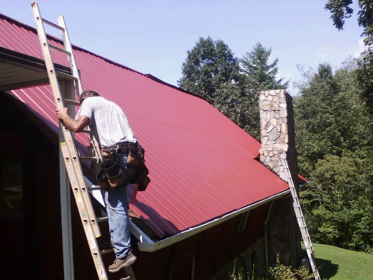 Roofer on a ladder beside a red metal roof and stone chimney — Soffit & Fascia Repair by Roofing Brothers in Albright